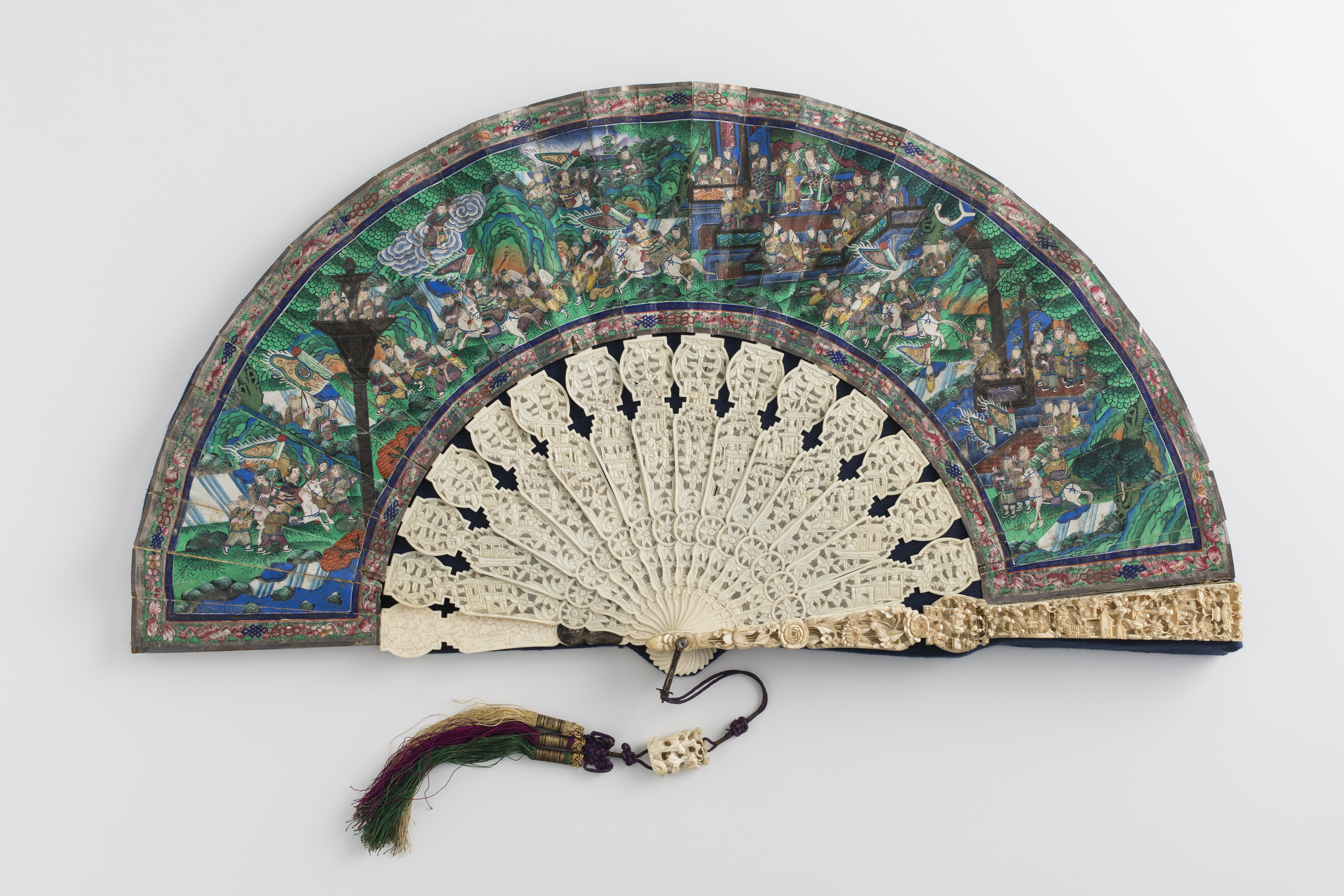 Chinese folding fan open on a grey background. The blades are intricately carved ivory and the painted paper surface (leaves) portrays a busy landscape, depicting what looks like a historic scene with many small brightly coloured figures.